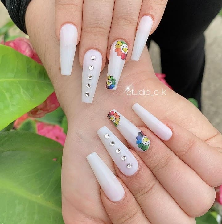 A closeup of a woman's fingernails with a white nail polish base that has rhinestones vertically and a rainbow sunflower art