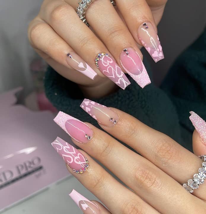 A closeup of a woman's fingernails with a nude and pink nail polish that has heart nail art, sugary glitter French tips and rhinestones