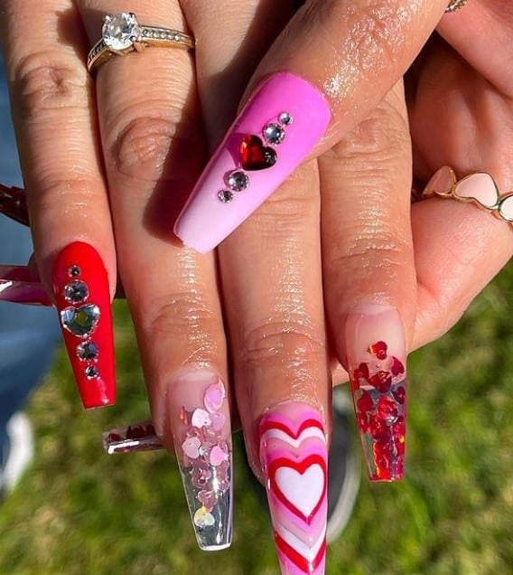 A closeup of a woman's fingernails with a combination of clear acrylic nails, pink ombré nails and red nail polish that has heart-shaped rhinestones and retro heart art on one nail