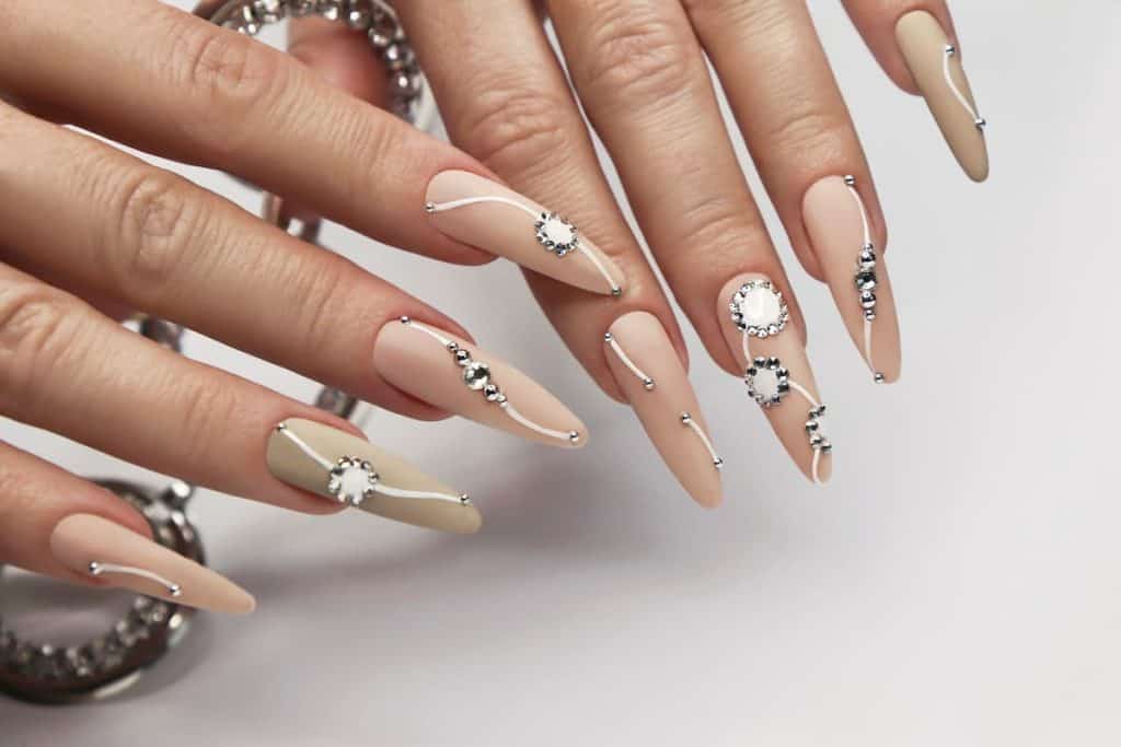 A closeup of a woman's fingernails with a matte-colored nude base that has curved white lines and the dots decorated with tiny rhinestones