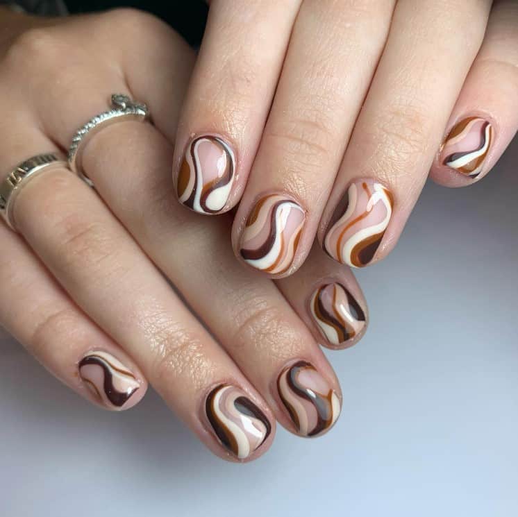 A closeup of a woman's fingernails with a nude nail polish base that has different shades of brown swirls with white streaks