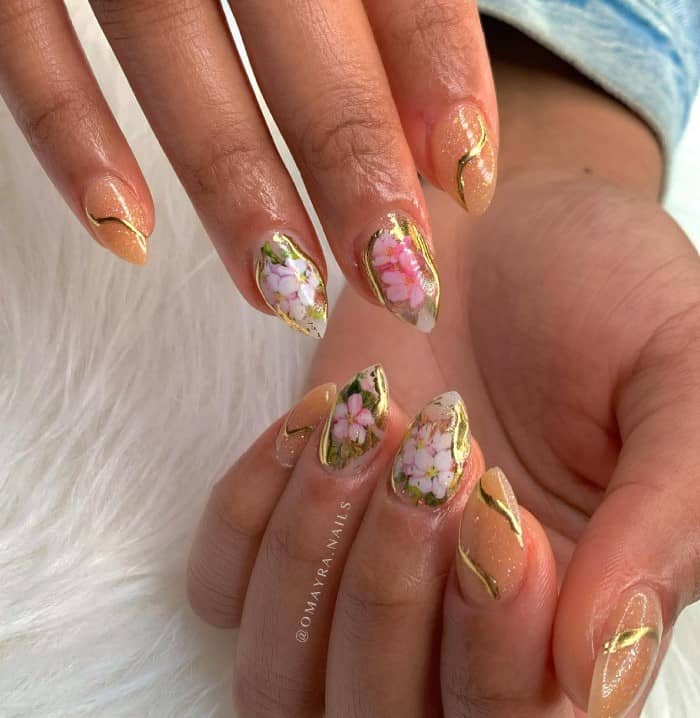 A woman's short stiletto fingernails with a nude nail polish that has  pressed flower decals and abstract wavy lines of gold