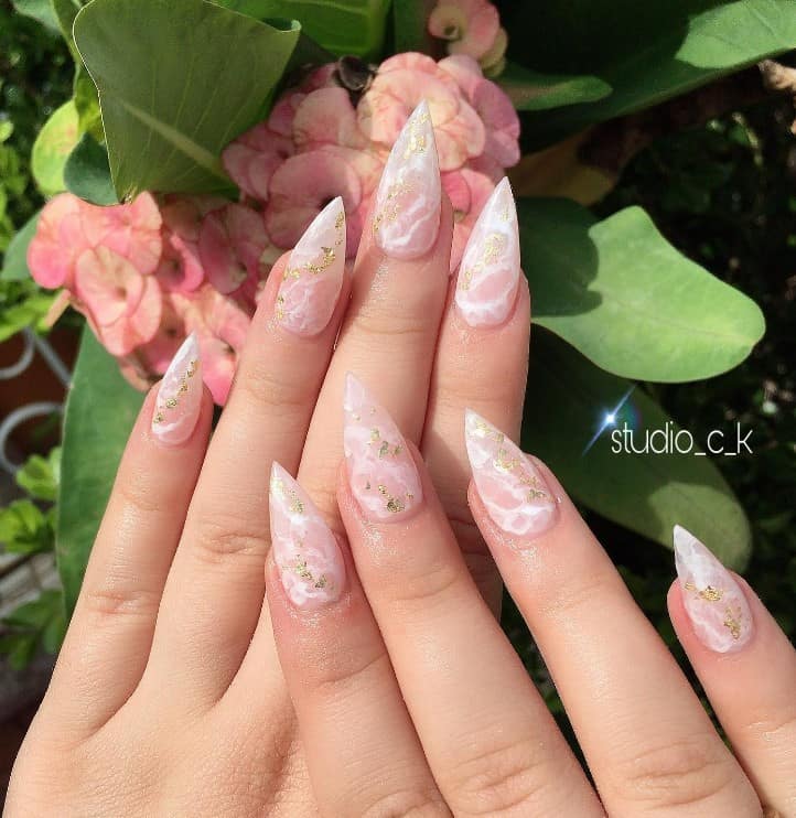 A closeup of a woman's fingernails with a combination of white and sheer pink polish that is a marble-like design that has gold flakes
