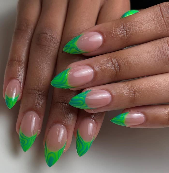 A closeup of a woman's pointy stiletto fingernails with a nude nail polish base that has green swirly French tips