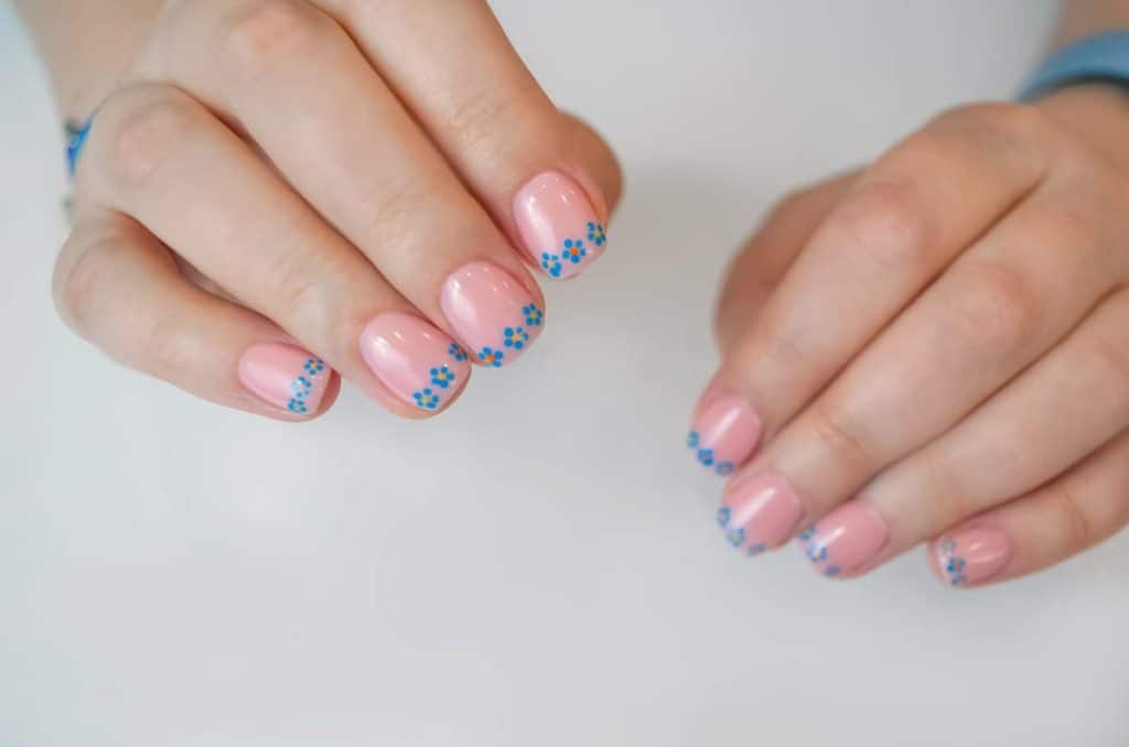 A woman's fingernails with a a light pink nail polish that has a line of blue flowers on nail tips
