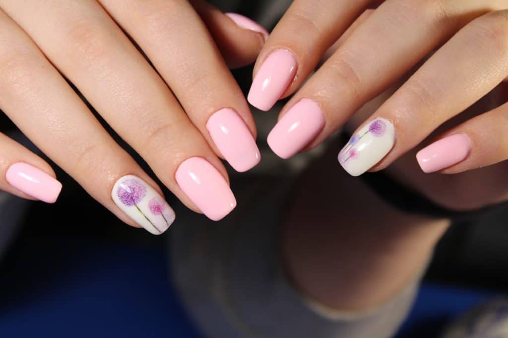 A closeup of a woman's fingernails with a combination of white and bubblegum pink nail polish that has dandelion art using blue, purple, and a darker shade of pink