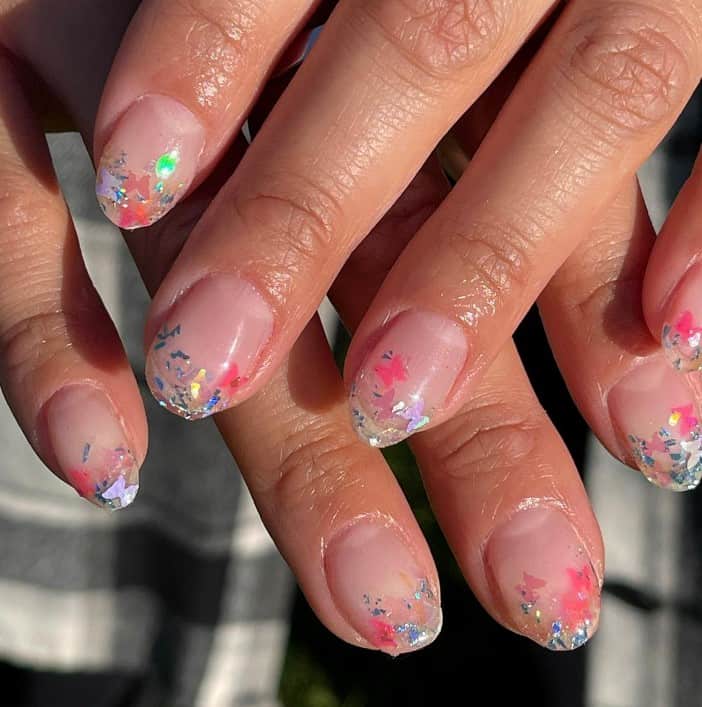 A closeup of a woman's fingernails with a nude nail polish base that has various colored flakes and butterflies on the nail tips 