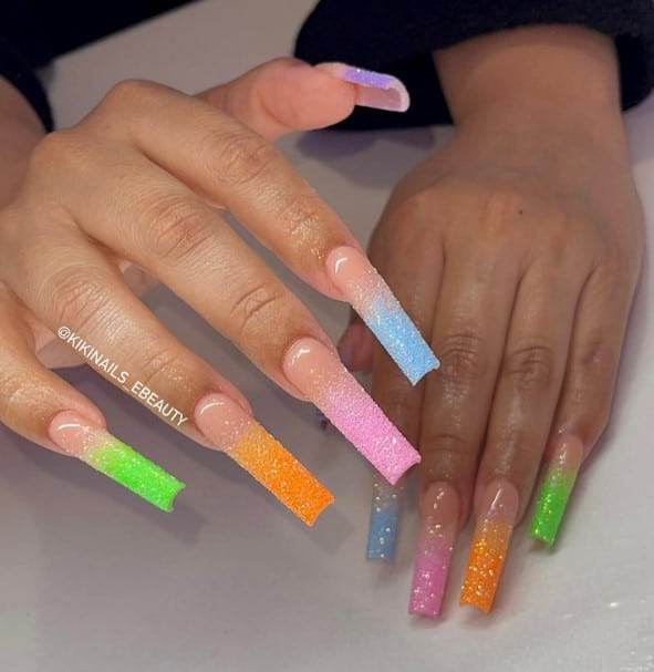 A closeup of a woman's fingernails with a peach nail polish base that has multicolored nail tips in bright glittery hues