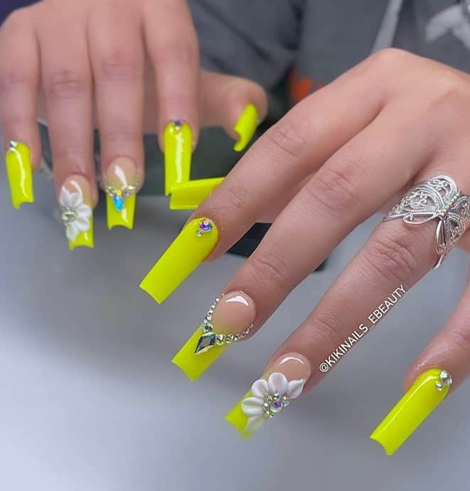 A closeup of a woman's fingernails with a neon yellow-green hue that has sparkling diamonds and rhinestones, and intricate 3D flowers