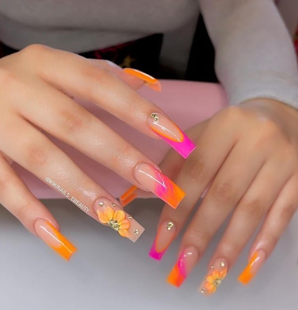A woman's fingernails with a nude nail polish base that has orange and pink hues, diamonds and 3D flowers 