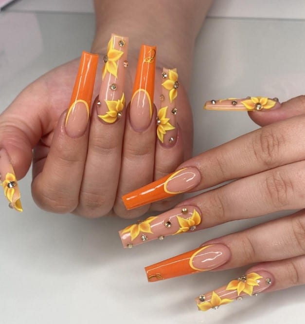 A closeup of a woman's fingernails with a nude nail polish base that has bright orange tips with yellow borders, yellow 3D flowers and rhinestones