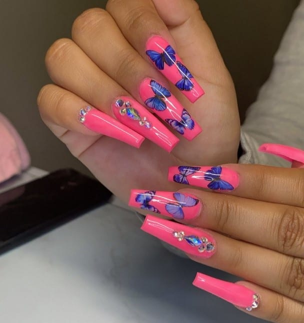 A closeup of a woman's hot pink base fingernails with midnight-blue butterflies that has Rhinestones and diamonds