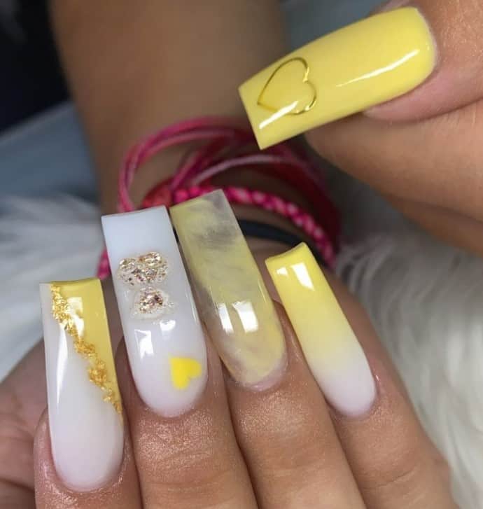 A closeup of a woman's thick acrylic fingernails with pristine white base and showcasing an array of varied yellow designs that has gold foils, hearts, ombré, and marble patterns