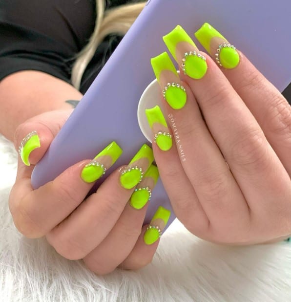 A closeup of a woman's fingernails with electrifying lime shade covers the base and tips that has neon base bordered with rhinestones
