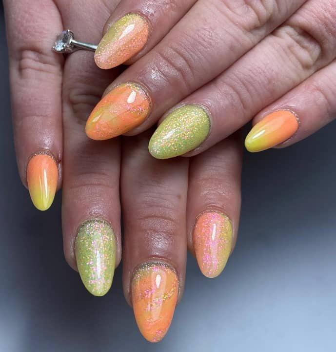 A closeup of a woman's fingernails with almond-shaped nails that has a punchy mix of light-green, yellow, and orange plus gold glitter
