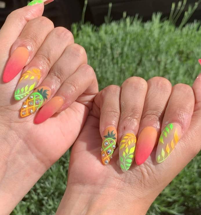 A woman's fingernails with an ombré of red to orange hues that has pineapples, pops of rhinestones, and orange, green, and yellow ombré fern silhouettes