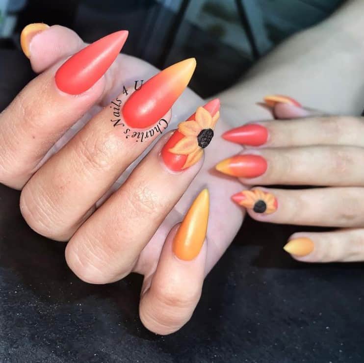 A closeup of a woman's stiletto fingernails with matte yellow and orange ombré that has add a 3D sunflower accent
