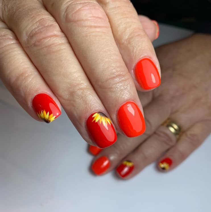 A closeup of a woman's fingernails with a glossy red-orange nail polish that has half-sunflower nail art on two nails