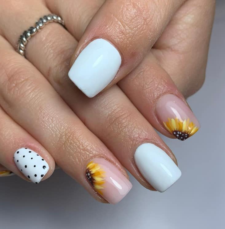 A closeup of a woman's fingernails with a combination of white and nude nail polish base that has one polka-dot nail and two sunflower nail accents on a clear base