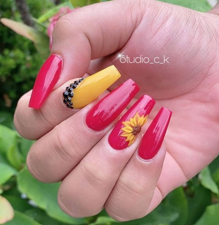 A closeup of a woman's fingernails with a mix of red and yellow nail polish base that has a 3D sunflower and the other with a black rhinestone-embellished nail on the yellow polish