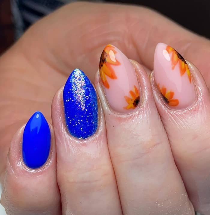 A closeup of a woman's fingernails with a combination of  a glossy and sparkling royal blue and nude nail polish base that has sunflowers nail design on nude polish 