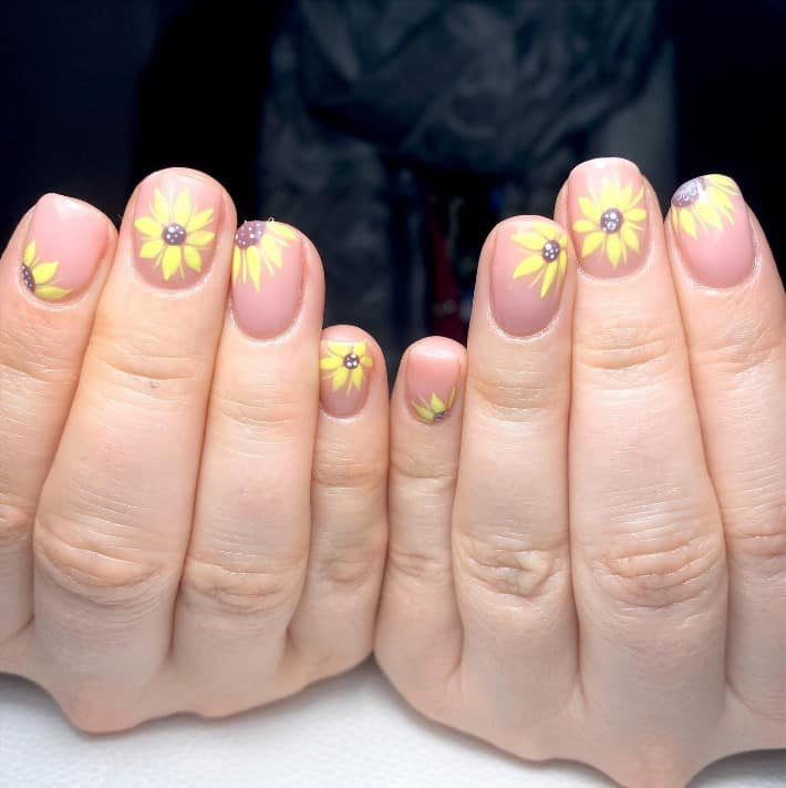 A closeup of a woman's fingernails with a nude nail polish that has different sunflower nail art with small gems in yellow polish