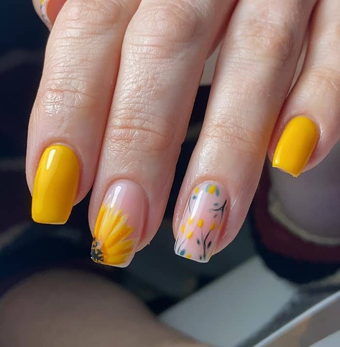 A closeup of a woman's fingernails with a mix of glossy nude and yellow nail polish base that has sunflower art and floral accents to clear nails