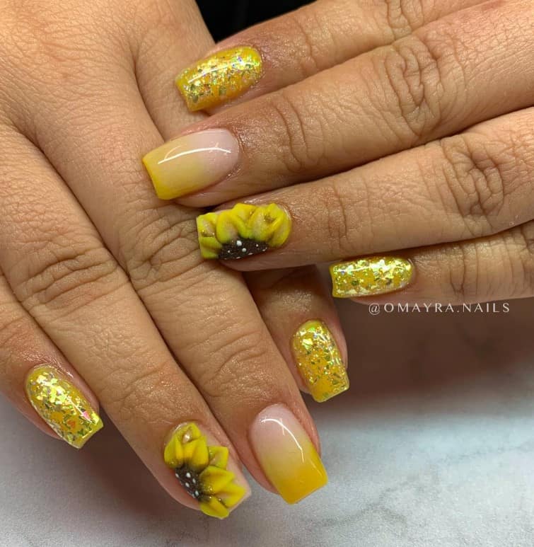A closeup of a woman's fingernails with gold glitter and yellow ombré nail polish base that has a 3D sunflower on one nail