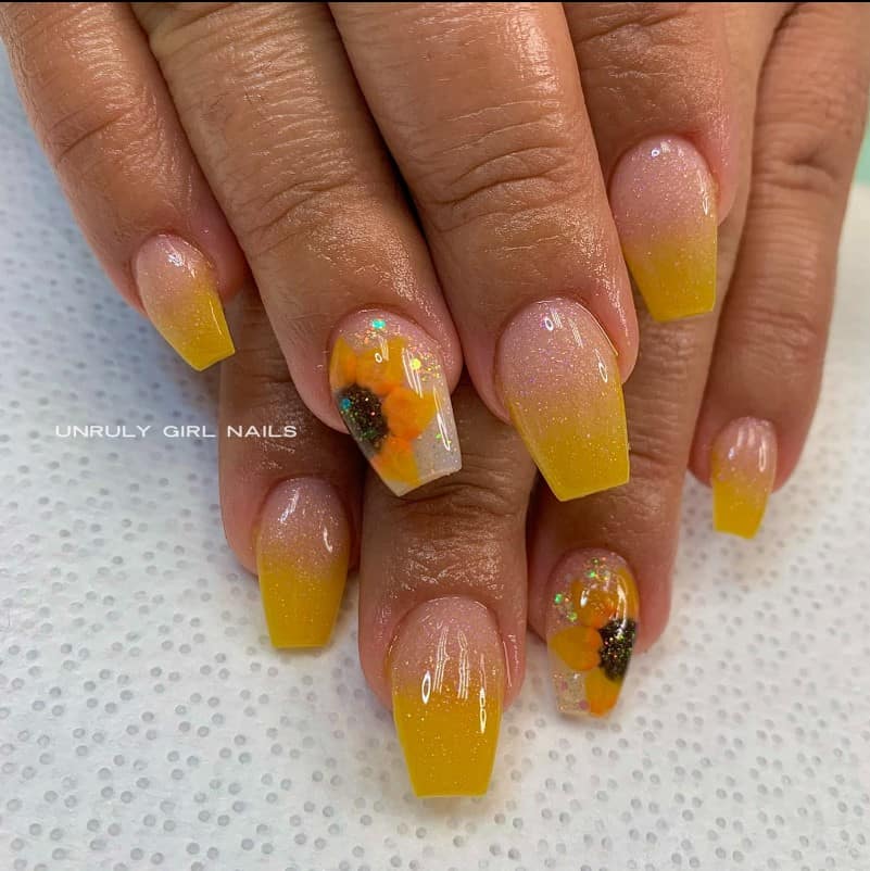 A closeup of a woman's fingernails with sparkly ombré manicure in clear and yellow polish that has a shimmering sunflower accent on select nails 