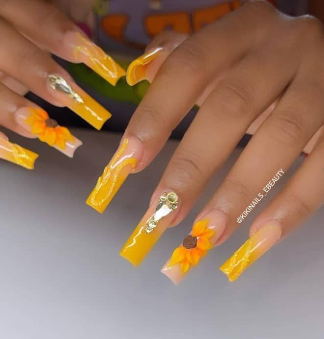 A woman's fingernails with an all-yellow ombré manicure that has glittery swirls, 3D flowers, and sparkling rhinestones