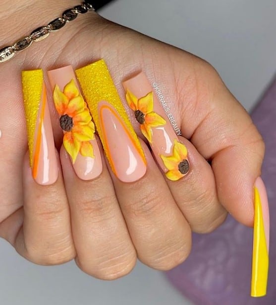 A closeup of a woman's fingernails with a nude nail polish base that has diagonal French tips in yellow polish and sugary glitter and different 3D sunflower designs
