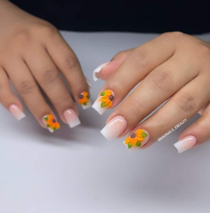 A woman's fingernails with a nude-and-white ombré color combination that has 3D sunflowers on the accent nails