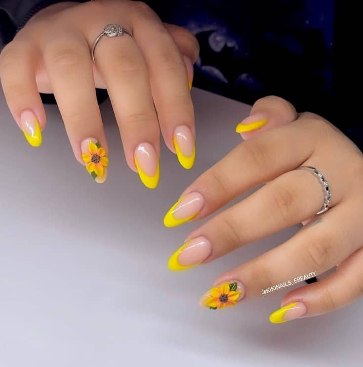 A woman's fingernails with a nude nail polish base that has yellow French tips and a 3D sunflower to one nail each hand 