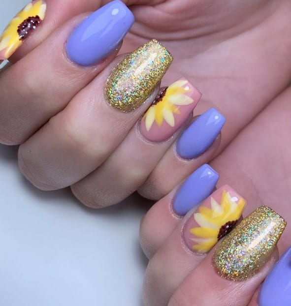 A closeup of a woman's fingernails with a combination of nude, purple and yellow glitter nail polish that has half-sunflower nail art on select nails 