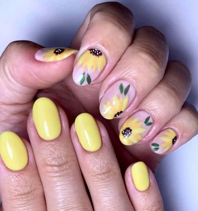 A closeup of a woman's fingernails with a nude nail polish base on one hand and yellow nail polish on the other that has sunflower nail art for accent on the nude nails 