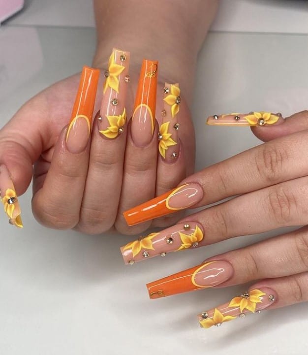 A closeup of a woman's fingernails with a glossy nude nail polish that has orange and yellow French tips, sunflower nail art and a few rhinestones