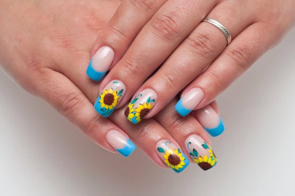 A woman's fingernails with a nude nail polish base that has sky blue French tips and detailed sunflower accents to them
