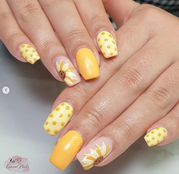 A closeup of a woman's fingernails with a combination of nude and yellow nail polish that has sunflower with white outlines on petals and polka dot designs 
