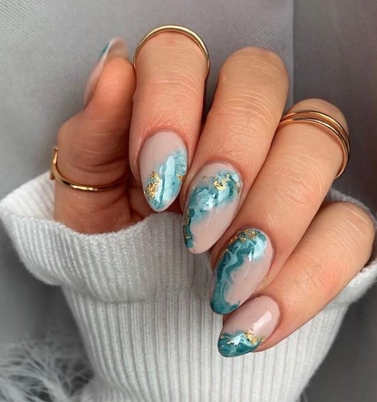 A closeup of a woman's fingernails with a nude nail polish that has cashmere-colored nails with turquoise ocean swirls and glimmering flecks of gold foil