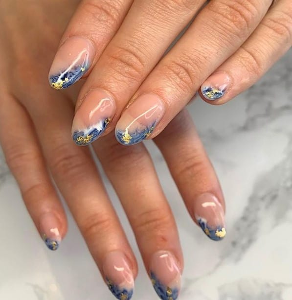 A closeup of a woman's fingernails with a nude nail polish that has blue-and-gold ocean wave tips