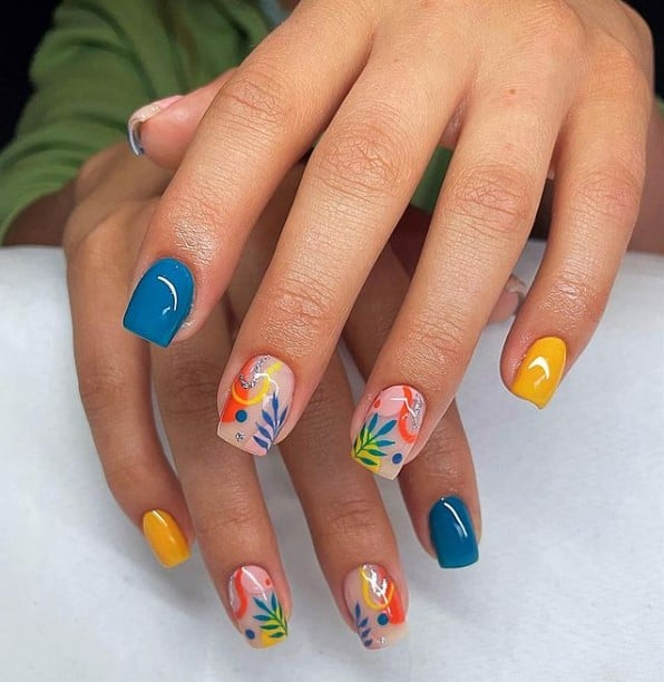 A closeup of a woman's fingernails with combination of turquoise and yellow nail polish that has abstract nail art and leaves nail designs