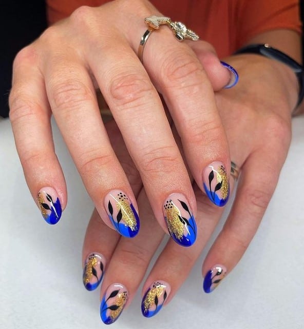 A closeup of a woman's fingernails with a nude nail polish that has cobalt blue tips, gold flakes, and black leaves