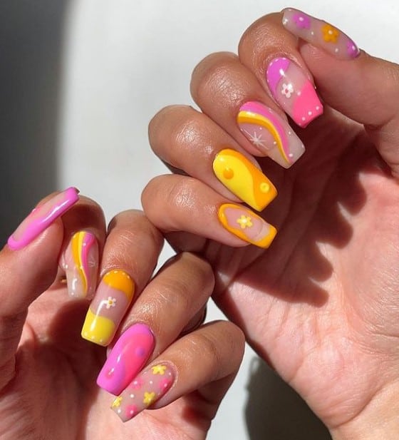 A closeup of a woman's fingernails with a nude nail polish that has hot pink, orange, and yellow in yin-and-yang and swirly patterns and adding floral accents
