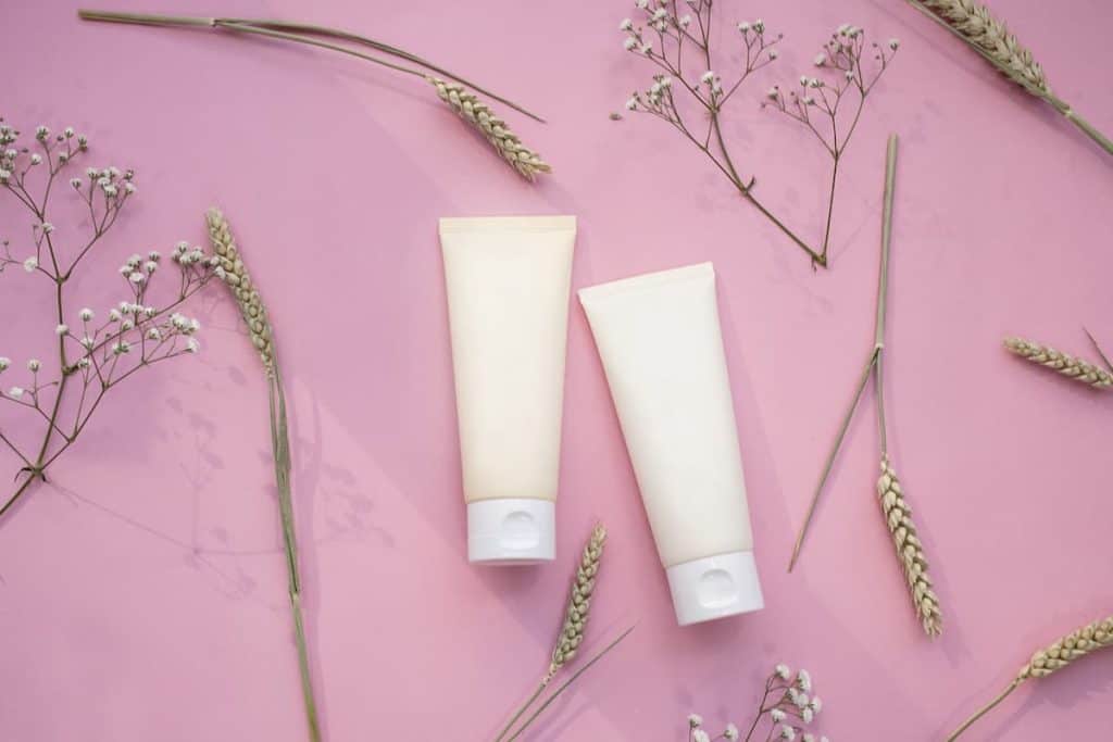 Two white cosmetic tubes in a flat lay with leaves and flowers isolated on a lilac background