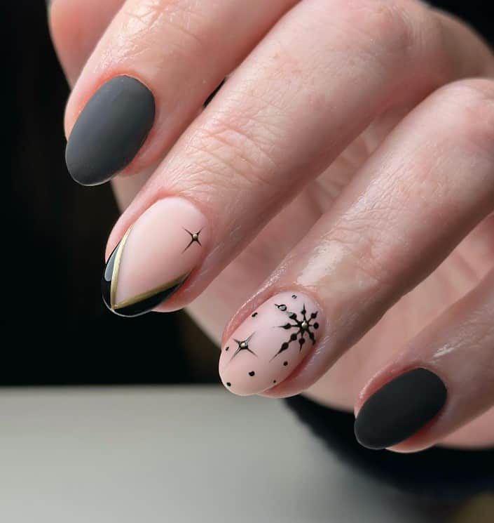 A closeup of a woman's fingernails with a combination of a matte black and nude nail polish that has black-and-gold V-tips along with black stars and snowflakes with gold gems