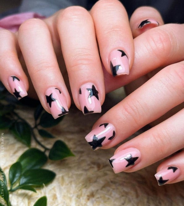 A closeup of a woman's square shaped fingernails with a glossy nude nail polish base that has black stars nail designs