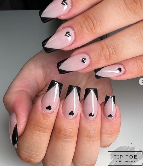 A closeup of a woman's fingernails with a nude nail polish base that has black chevron tips and little hearts on the cuticles