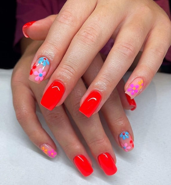 A closeup of a woman's long nails with nude pink and bright red nail polish that has multicolored vibrant flowers on nude nails 
