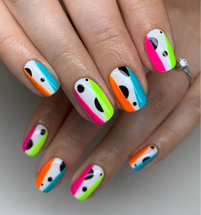 A closeup of a woman's short nails with white nail polish base that has irregular lines in mismatched colors on the sides and asymmetrical black circles nail designs