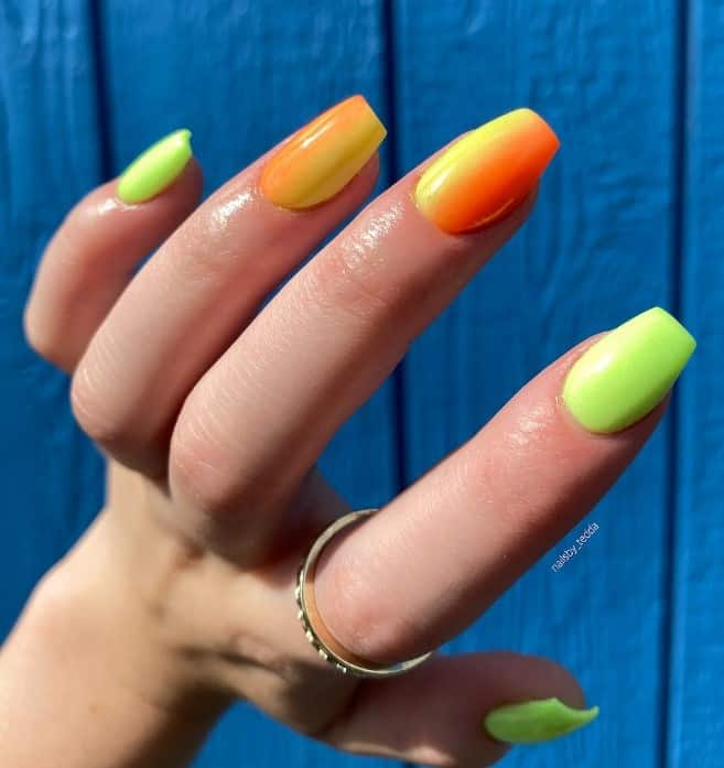 A closeup of a woman's mid-length coffin nails with bright green and shades of orange and yellow blend vertically on the accent nails in an ombre effect